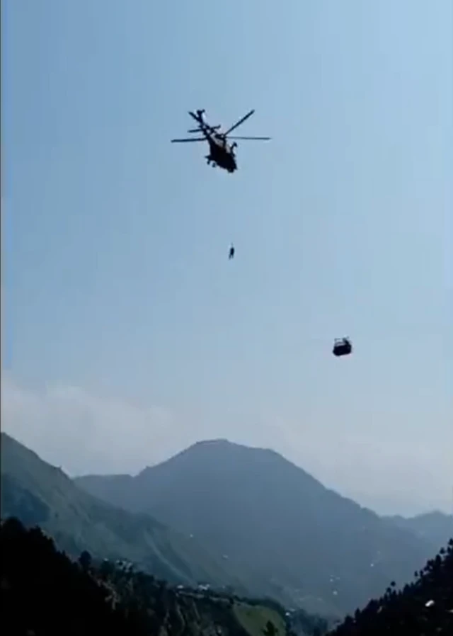 A man descending from a helicopter towards a suspended cable car, with mountains in the background