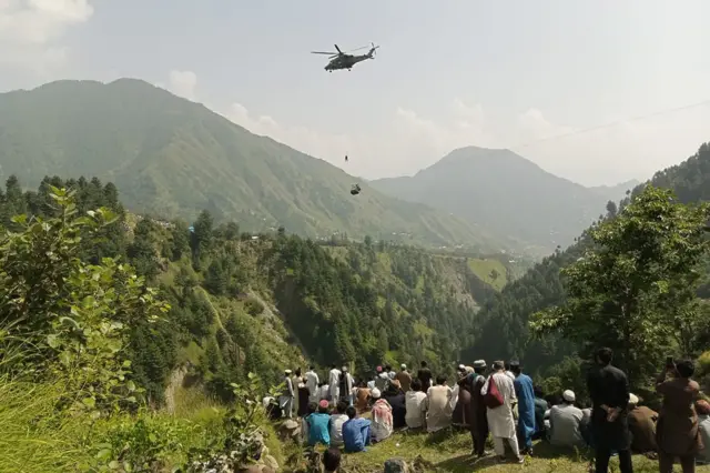 People on a mountainside watch as an army helicopter hovers over a suspended chairlift in the mountains