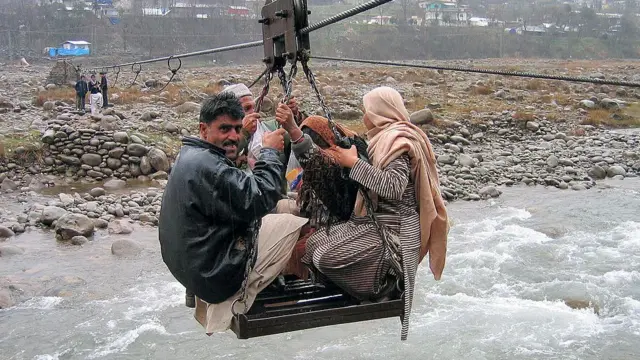 A group of people travel on a cable car across a body of water, holding onto nothing other than chains