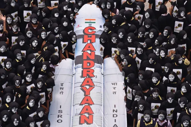 A group of students with faces painted like the moon gather around a model of the Chandrayaan-3 rocket