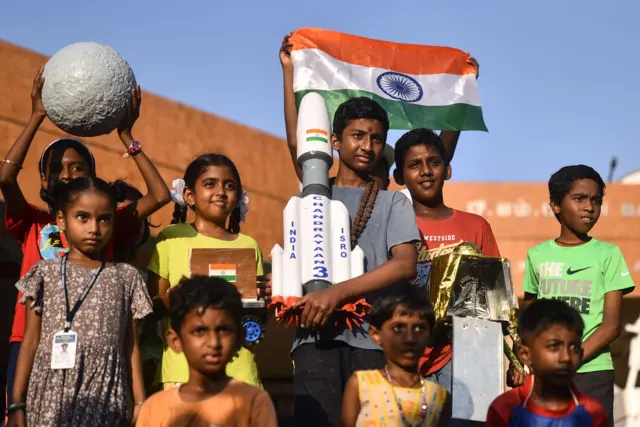 A group of children holding the Indian flag and models of the moon and the Chandrayaan-3 spacecraft