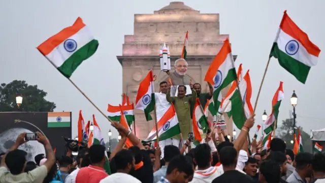 A crowd in the street waves Indian flags and a cut-out of Modi