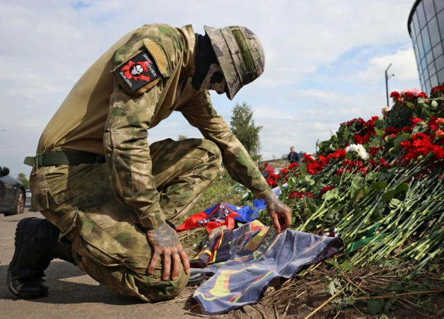 A man in Wagner uniform kneels in front of a bed of roses in tribute to Prigozhin