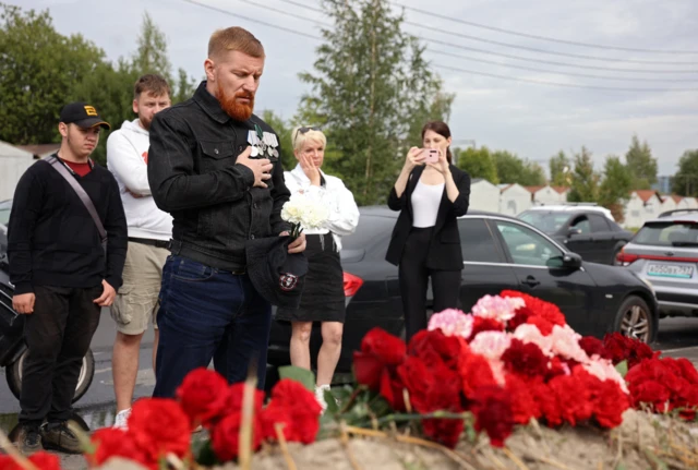 A man with his hand on his heart in front of a bed of roses paying tribute to Yevgeny Prigozhin