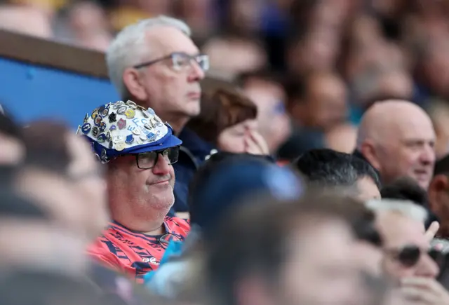 An everton fan sits grumpily in his pin-badge adorned flat cap.