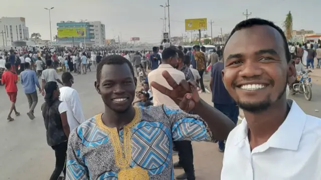 Two Sudanese men smile for selfie in a busy outdoor spot.