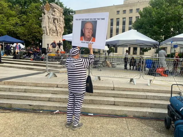 A demonstrator in a mock black and white striped prison outfit holds a up a sign of an older Trump, with the caption "you do the crime you pay the time, your time is up"