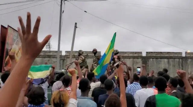 A crowd gathers as soldiers drive past them, waving Gabonese flags