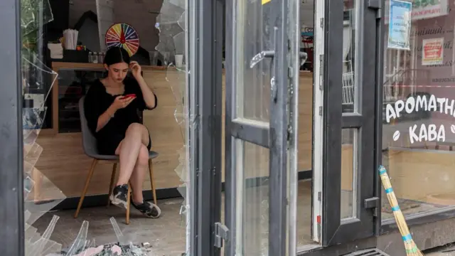 A woman sits on a chair, framed by the jagged remains of a glass door