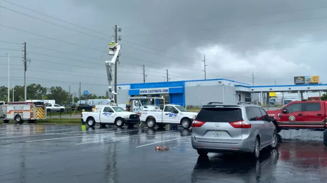 A worker is in a cherry-picker, raised up to some power lines. The sky above is dark grey