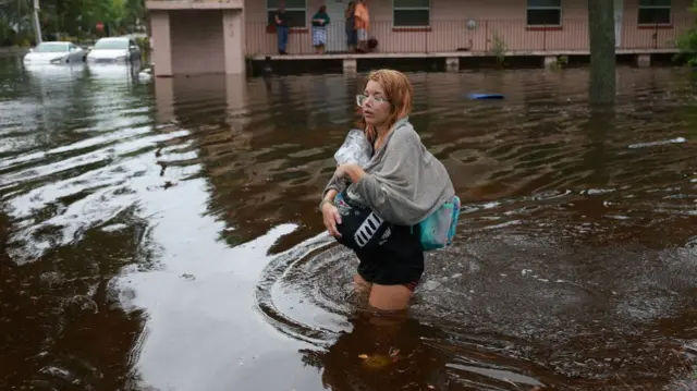 A woman carries bags and a towel as she walks through floodwaters up to her thigh