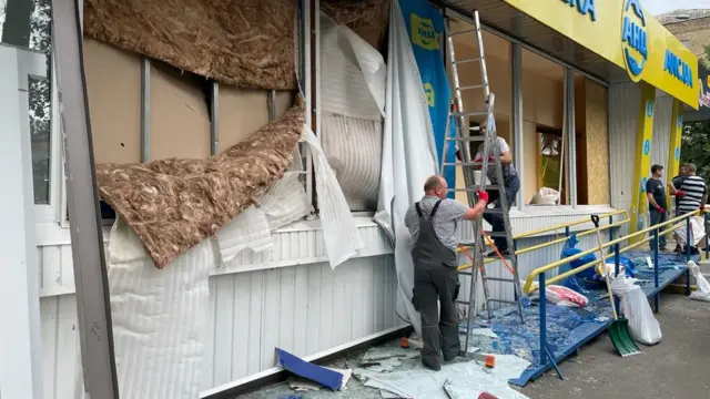 A damaged shop front and pharmacy with shattered glass on the floor and insulation hanging from the wall