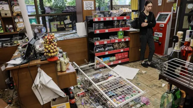 A young woman stands in a shop among shattered glass and toppled shelves