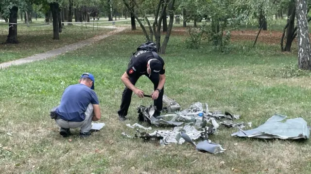 Two men examine shards of warped metal on a grassy area near a gravel path