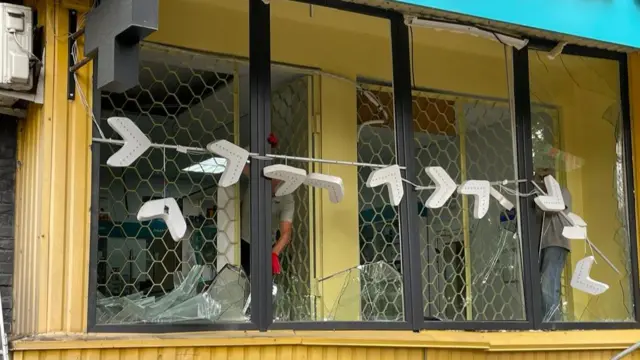 Broken windows carry shards of broken glass, with red and white tape across the front of the yellow building