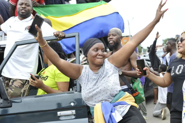 A group of people - some sitting in a vehicle - wave flags and appear to celebrate a coup in Gabon