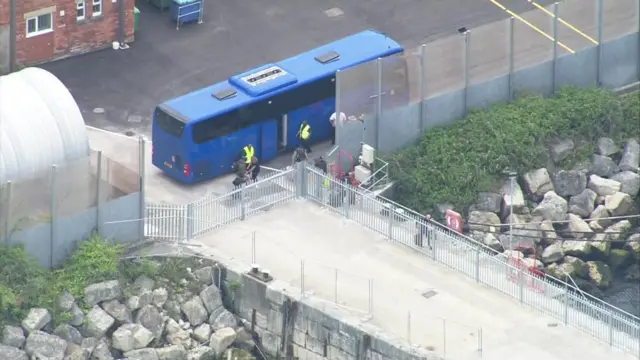 Blue coach parked outside entrance to Portland harbour, with several people walking away from it towards the Bibby Stockholm