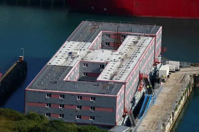 The Bibby Stockholm barge, a three storey rectangle barge moored in Portland