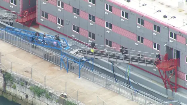 Six people boarding the ramp of the Bibby Stockholm, they are all carrying bags