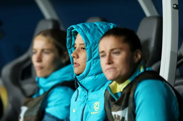 Sam Kerr sits glum with her hood up on the bench.