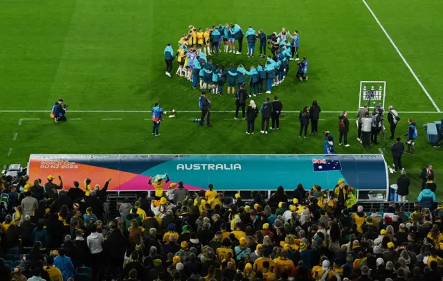 Australia players huddle together on the pitch with fans watching on from the stands.