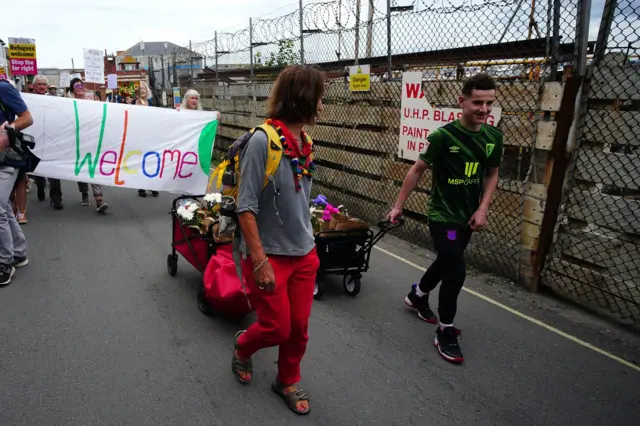 Two people carry trailers of welcome packs as people behind them hold a banner with a peace sign sayin welcome