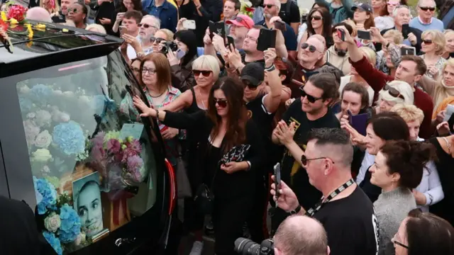 Crowds gather outside the late singer's home and one woman touches the hearse as it goes past