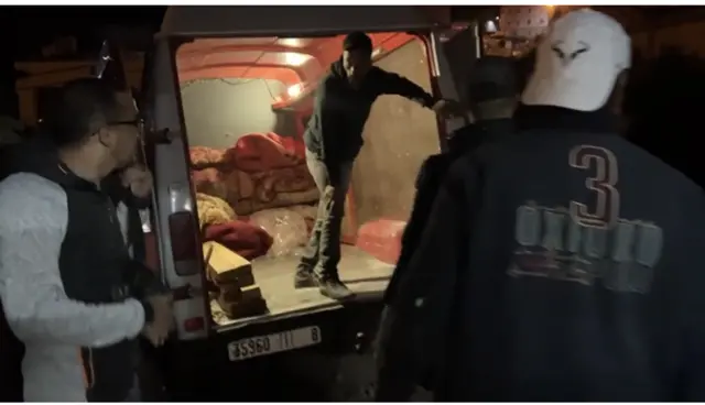 A man stands in the back of a van, speaking to two other men