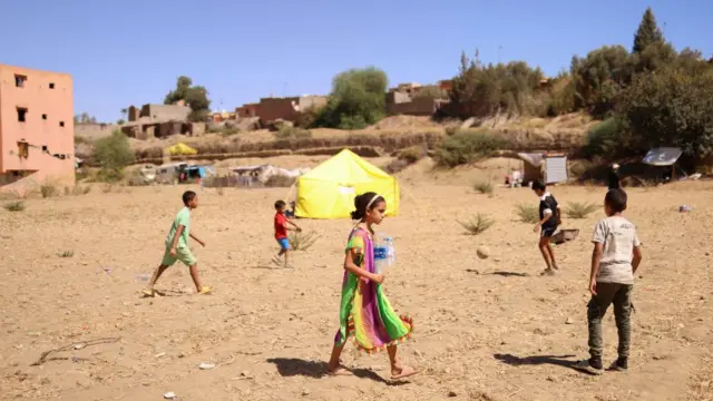 Children playing football on a dirt field next to a bright yellow relief tent