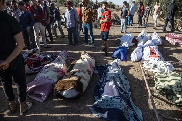 Mourners stand next to rows of bodies wrapped in rugs, laid out in a row