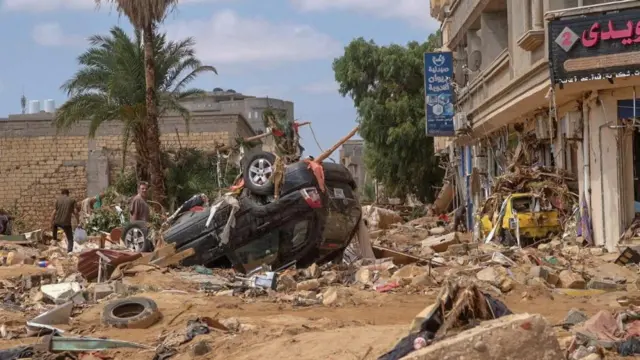 A car is upturned in the middle of a street littered with debris and caked with mud