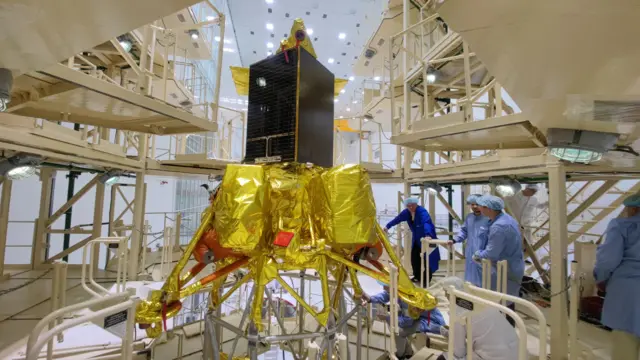 A group of scientists and technicians in blue working on a craft, covered in golden foil, that was meant to land on the moon