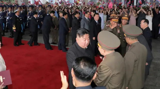 Kim Jong Un shakes hands with a member of the military on a red carpet. Various other suited and uniformed men accompany him