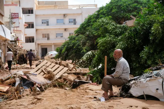 A man sits on a ruined car in front of debris and a fallen tree in a residential area