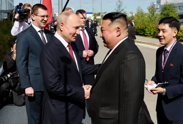 Vladimir Putin and Kim Jong Un shaking hands, surrounded by officials from both countries