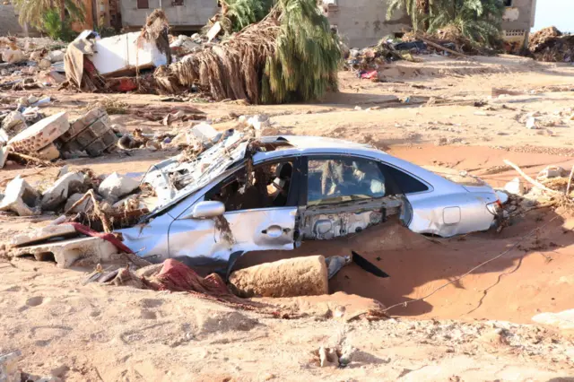 A destroyed car is half-buried under sand in Derna