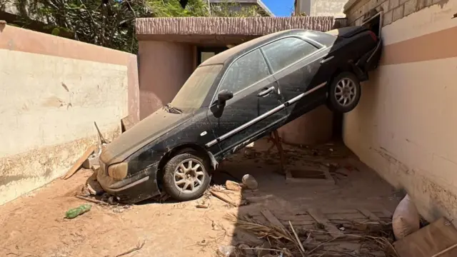 A car swept up by floods leans diagonally against a wall