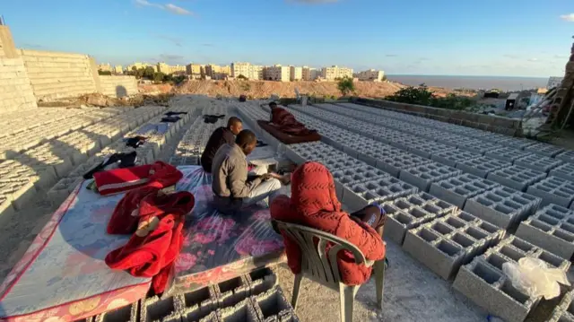 Men sit on mattresses placed outisde on piles of unusued breeze blocks