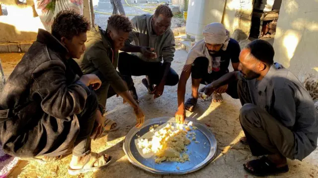 Five men squatting down next to a large platter of rice, sharing the food.