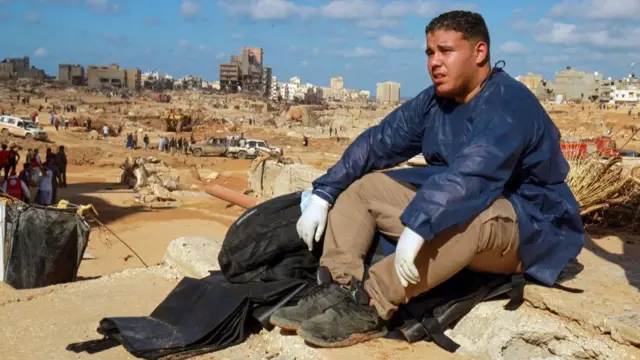 A man in scrubs sits on the muddy ground looking exhausted, Derna