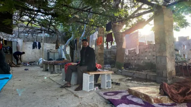 A man sits on a makeshift table made of a wooden board and 4 bricks. Clothes are hanging out to dry above him on strings hung between temporary structures.