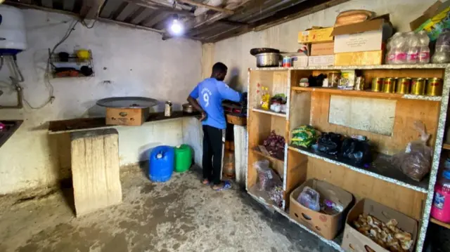 Man cooks in a makeshift kitchen, with a bare lightbulb hanging and open shelves storing basic ingredients like rice, onions and cooking oil,