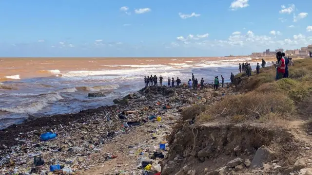 A crowd of people stand on a beach littered with debris in Derna 13  September