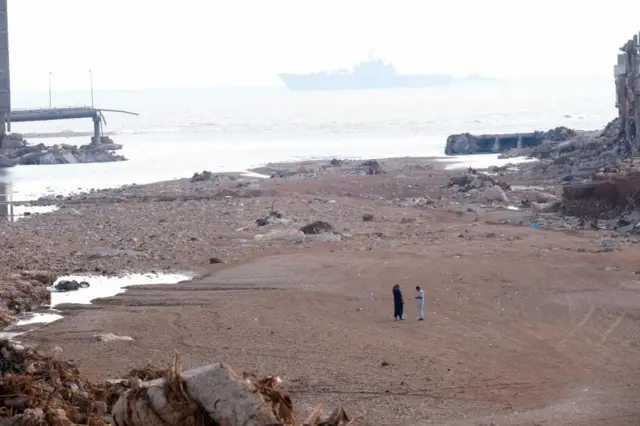 Two men stand on the beach after the devastating floods in Derna, with  rubble all around and a ship visible in the background