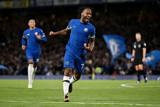 Raheem Sterling celebrates scoring versus Luton witha  flag waving in the background.