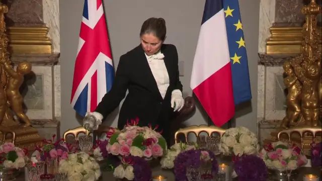 Waiter pours drinks ahead into glass on table filled with flowers.