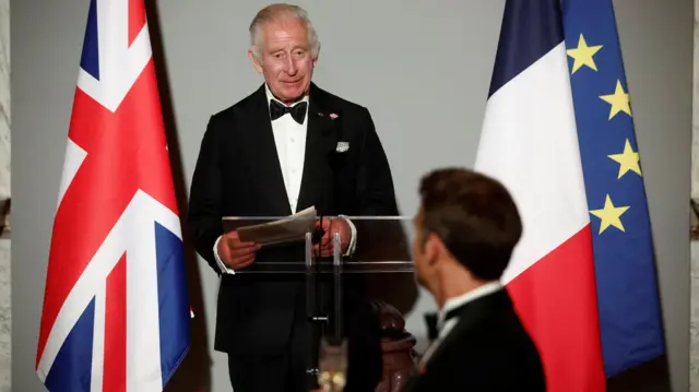 King Charles stands behind podium with Emmanuel Macron in the foreground, looking at one another