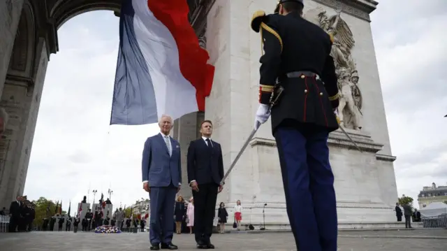 The King andf Macron stand together in front of the Arc de Triomphe, facing a soldier