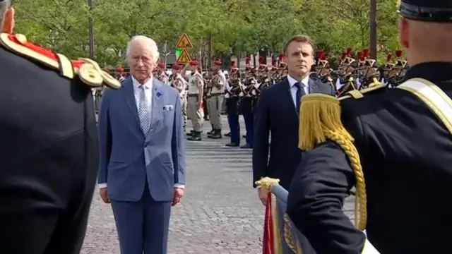 Charles and Macron stand listening to the national anthem