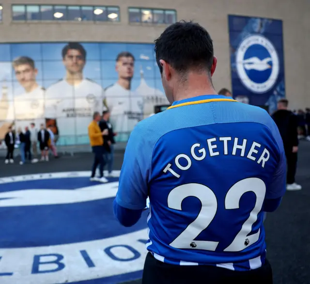 A Brighton fan stands infront of the ground wearing a shirt adorned with 'Together 22' on the back.